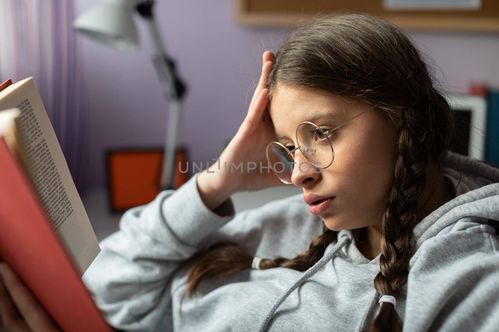 A schoolgirl is reading a highly engaging adventure book. The brunette has her glasses on and her hair styled in two braids. Book lover.