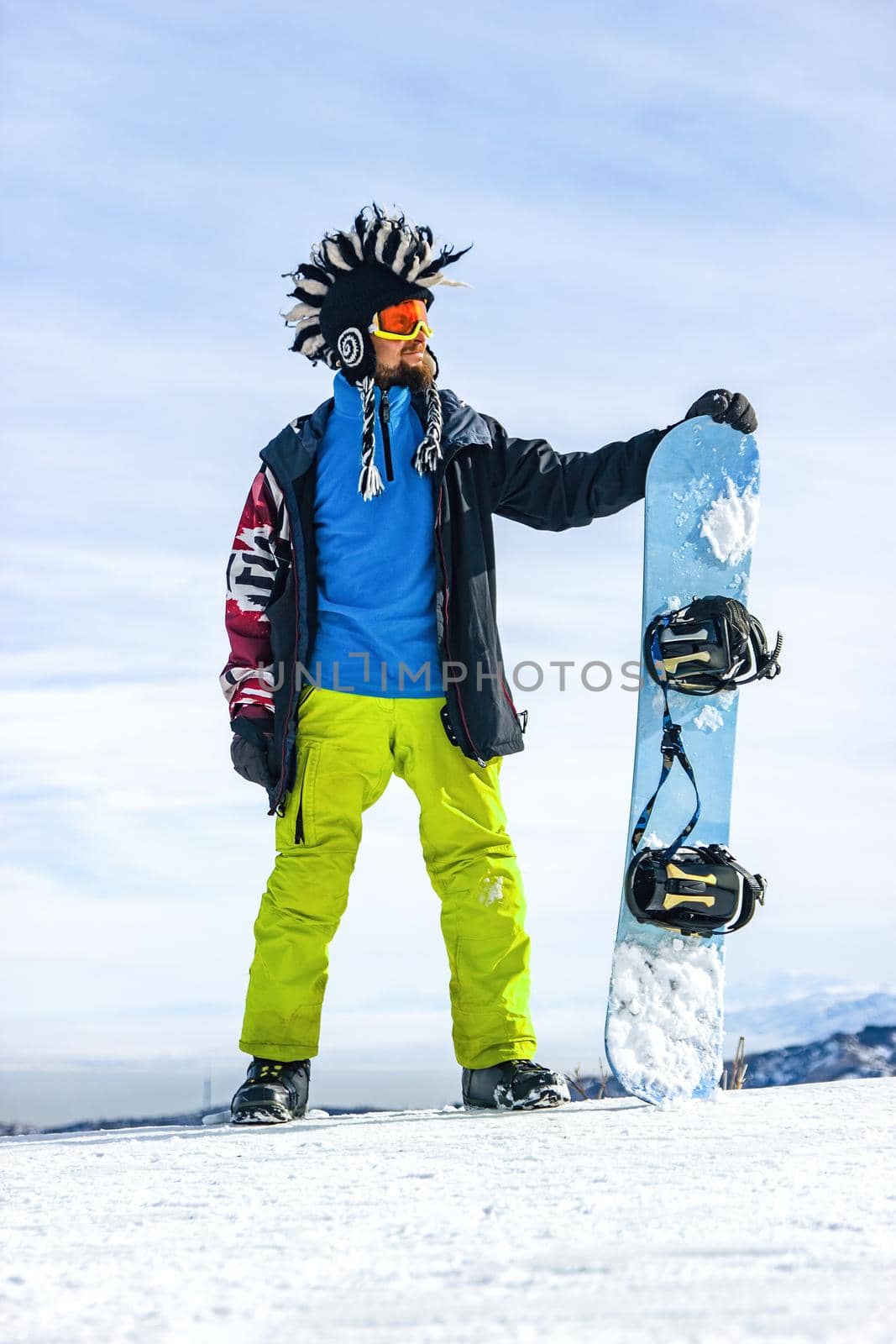 Bearded happy snowboarder in a ski mask with goggles and a fur big mohawk hat on a background of a sky and winter snowy mountains