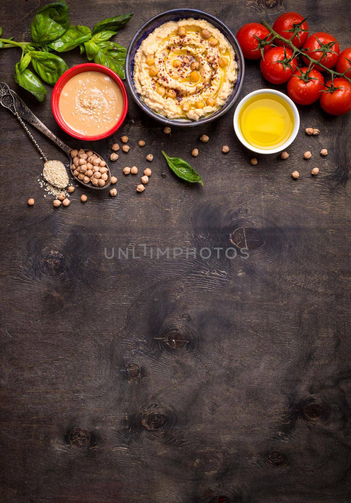 Bowl with hummus, chickpea, tahini, olive oil, sesame seeds, cherry tomatoes and herbs on dark rustic wooden background. Space for text. Food frame. Middle eastern cuisine. Top view. Hummus background