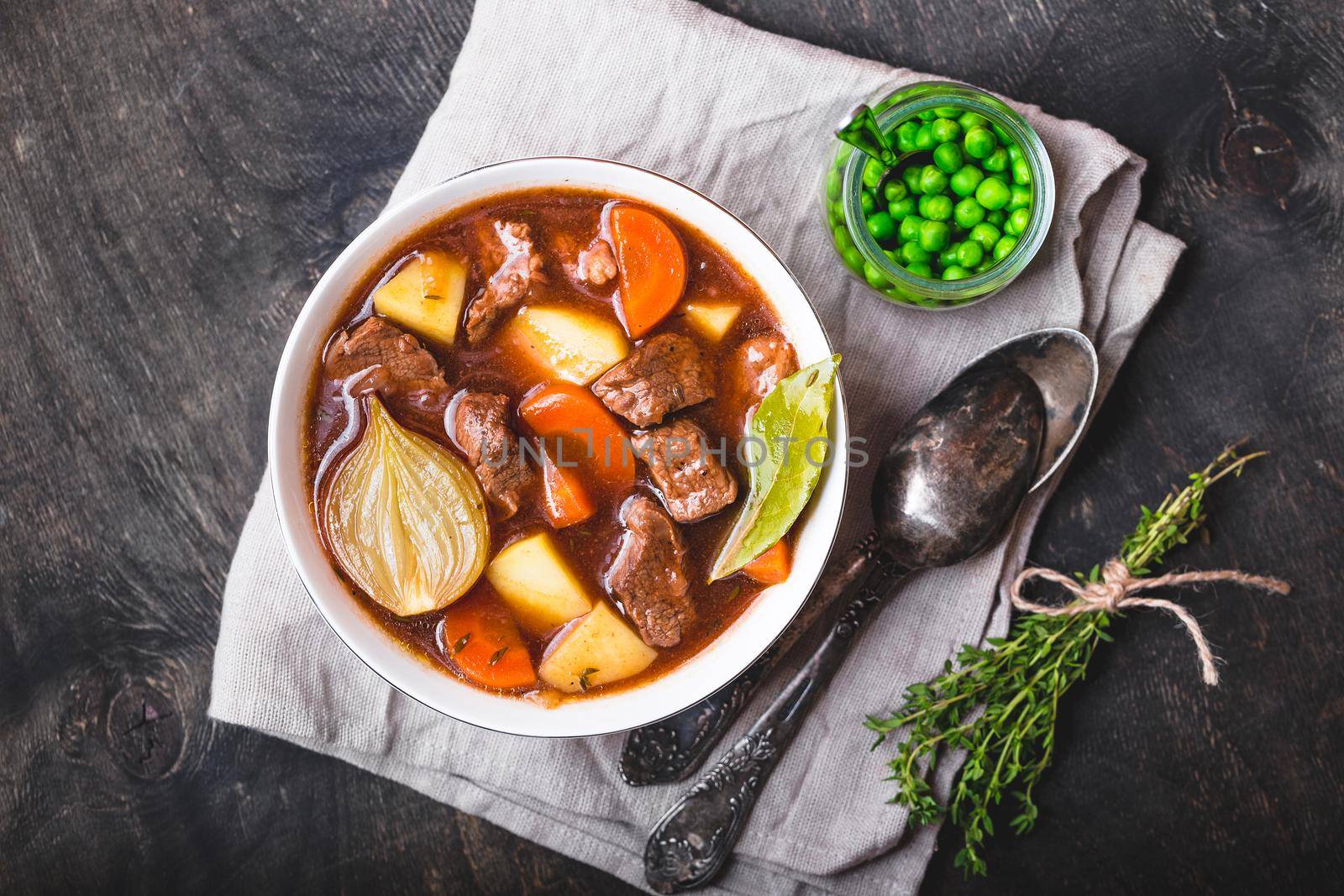 Meat stew with beef, potato, carrot, onion, spices, green peas. Slow cooked meat stew, bowl, wooden background. Hot autumn/winter dish. Closeup. Top view. Comfort food. Homemade soup/ragout/casserole