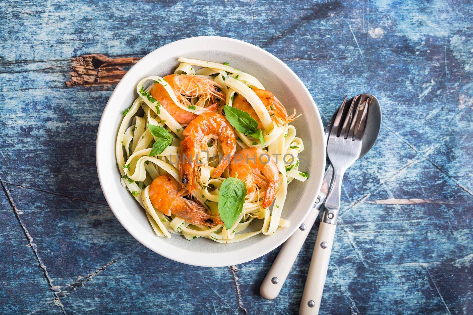 Homemade pasta with fried shrimps and basil on rustic wooden blue background. Pasta with seafood in plate. Top view. Close-up. Mediterranean cuisine. Pasta for dinner or lunch. Linguine with prawns