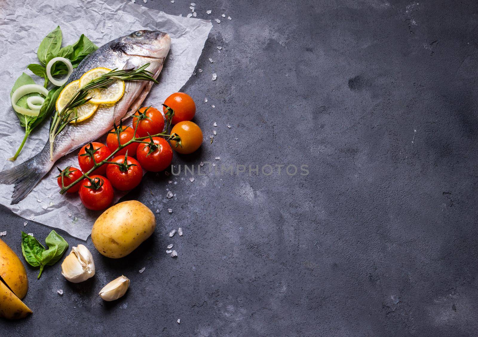 Raw fish with fresh ingredients ready to cook. Fish, lemon, herbs, potato, tomatoes. Ingredients for cooking on dark rustic background. Space for text. Diet and healthy food. Fish background. Top view