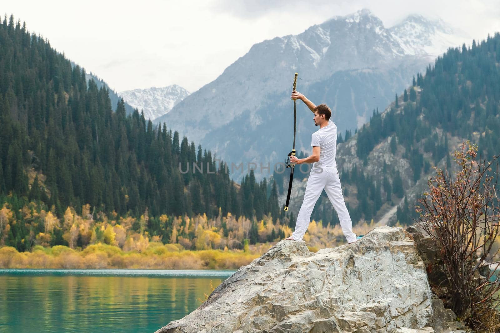 A young man stands on a large stone and holds a Japanese sword in his hands.