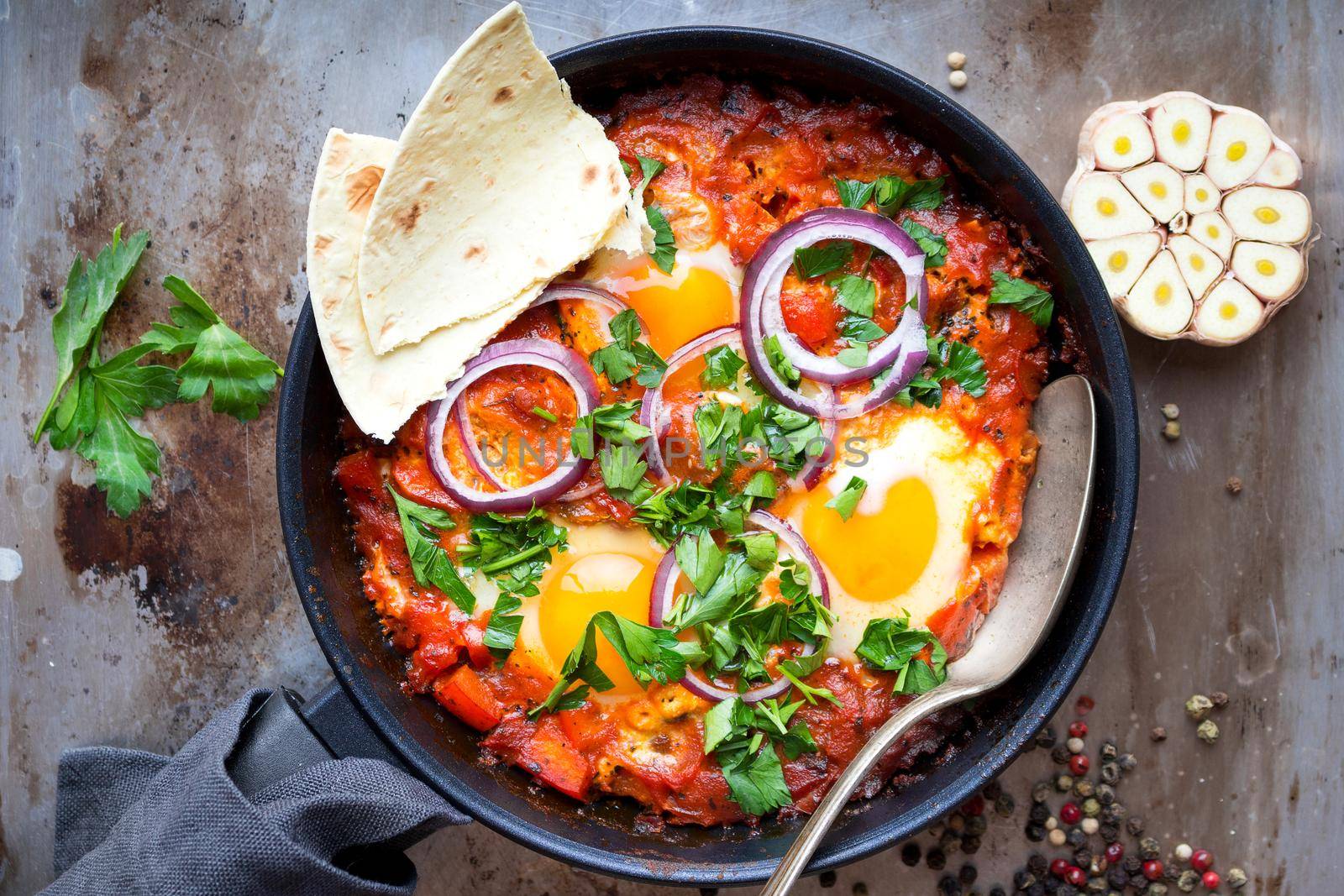 Shakshuka with pita bread in a pan. Middle eastern traditional dish. Fried eggs with tomatoes, bell pepper, vegetables and herbs. Shakshouka on a table. Top view. Sunny side up eggs. From above
