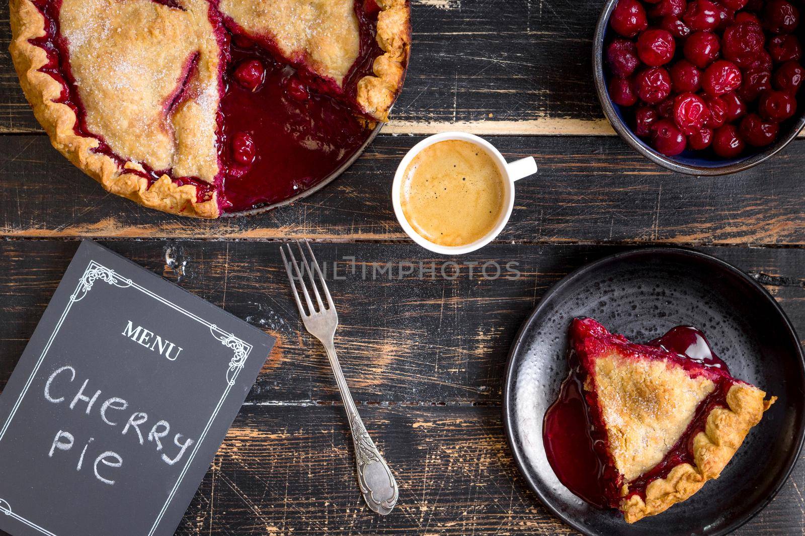 Homemade sliced cherry pie with flaky crust, cup of coffee, bowl with cherries and menu chalkboard on the black wooden table. Top view