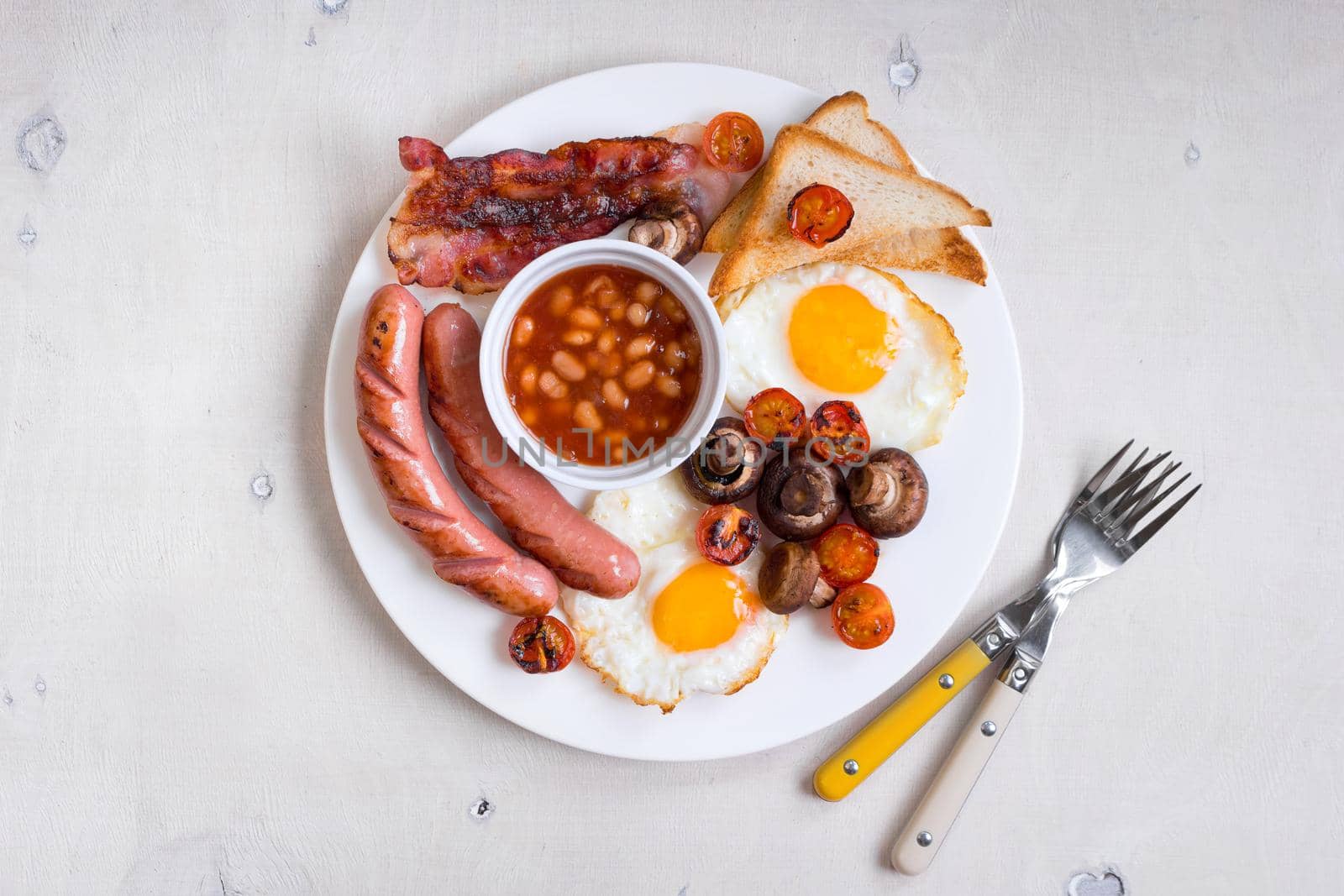 Full english breakfast with fried eggs, tomatoes, sausages, bacon, mushrooms, toasts and beans. Breakfast on a white plate with forks on the white wooden table. Top view