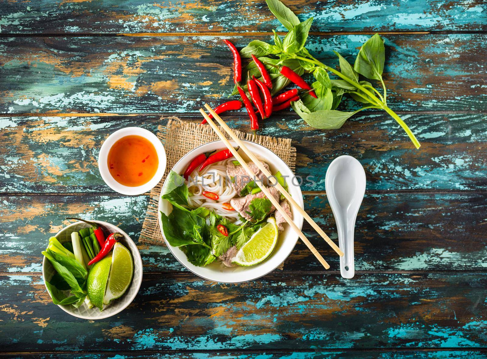 Traditional Vietnamese soup Pho bo with herbs, meat, rice noodles, broth. Pho bo in bowl with chopsticks, spoon. Top view. Asian soup Pho bo, wooden table background. Vietnamese authentic tasty soup