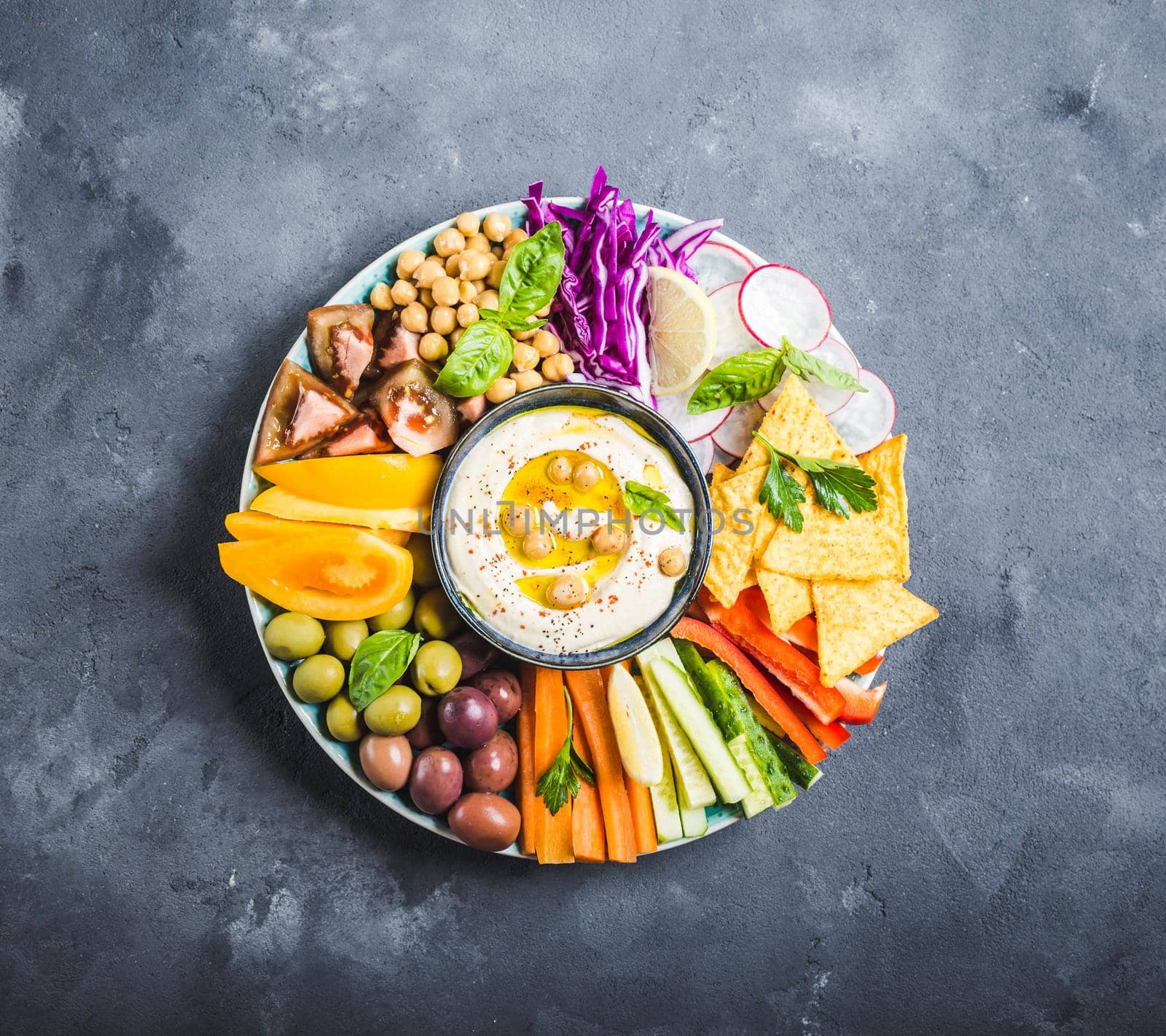 Hummus platter with assorted snacks. Hummus in bowl, vegetables sticks, chickpeas, olives, pita chips. Plate with Middle Eastern/Mediterranean meze. Party/finger food. Top view. Vegetables, hummus dip