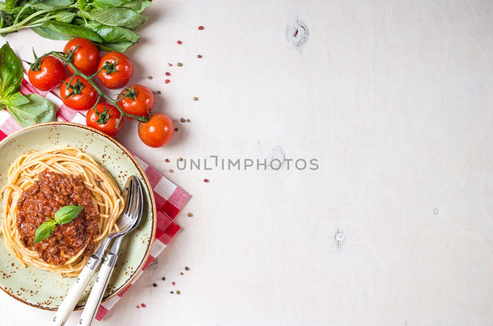 Italian pasta bolognese. Spaghetti with meat and tomato sauce in a plate with Italian tablecloth on a wooden white background. With fresh cherry tomatoes, basil. Food frame. Space for text. Top view