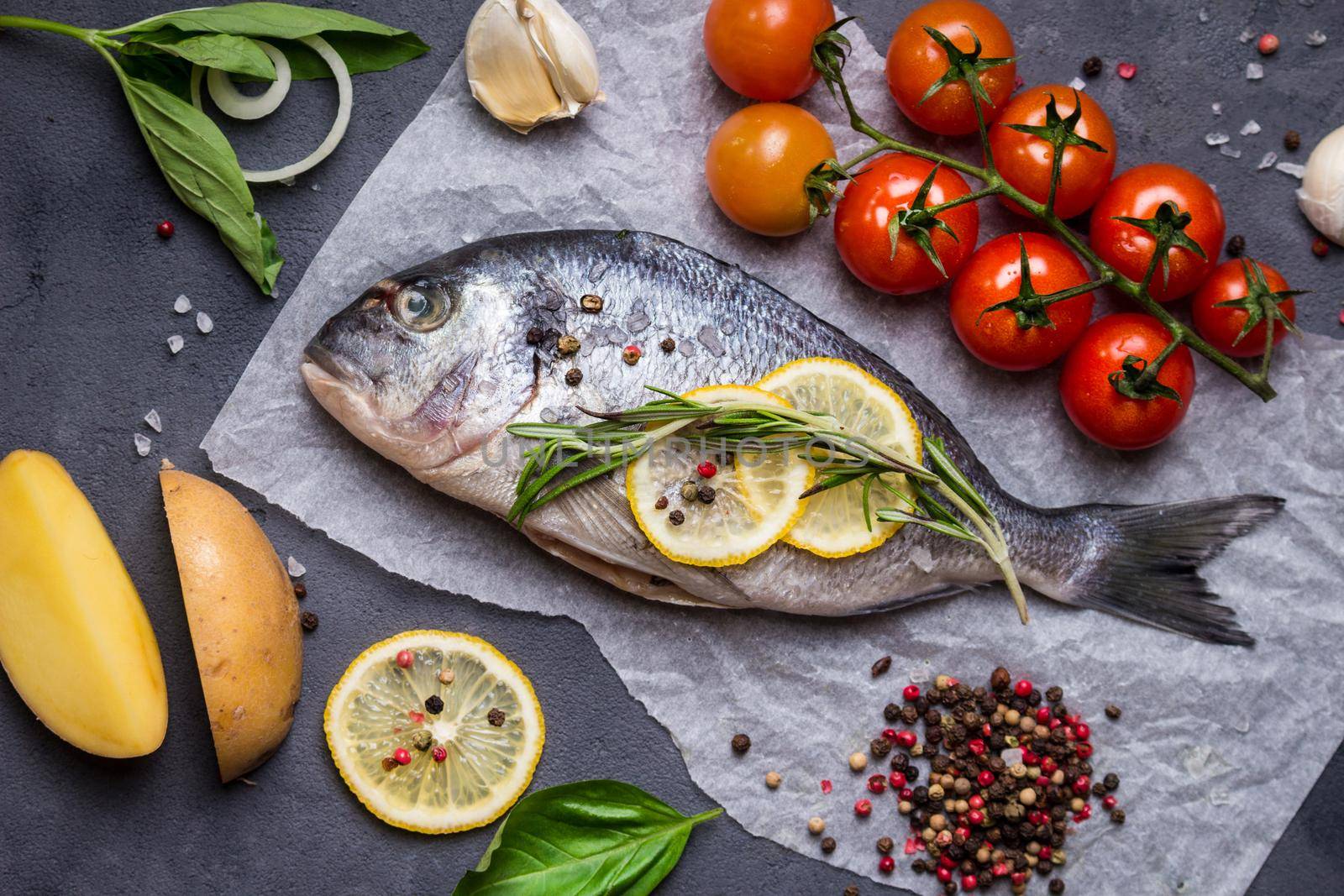 Raw whole fish with fresh ingredients ready to cook. Sea bream, lemon, herbs, potato, tomatoes, garlic, spices. Ingredients for cooking on dark rustic background. Diet and healthy food. Top view