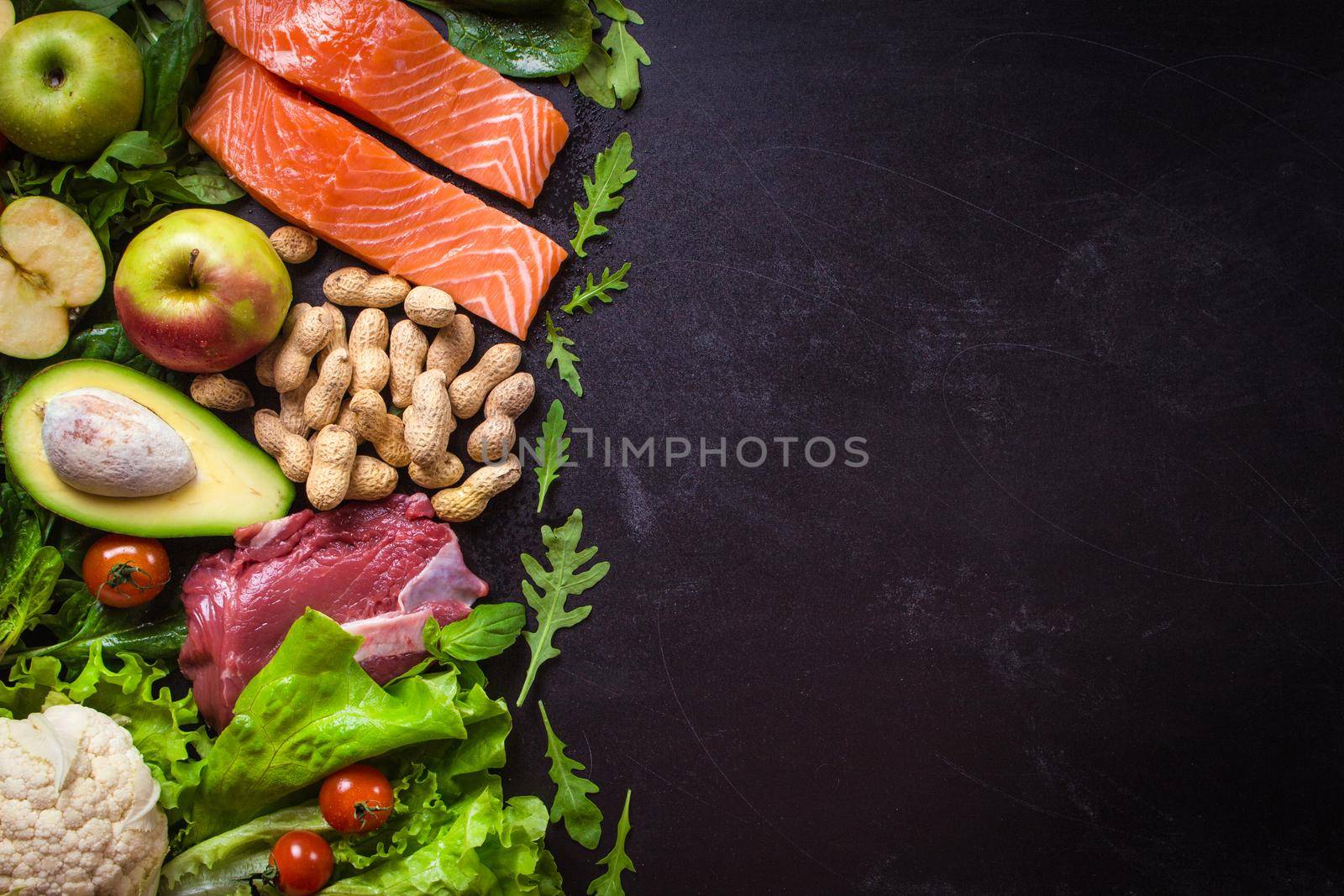 Fresh vegetables, fruits, fish, meat, nuts on black chalk board background. Сauliflower, avocado, apples, tomatoes, salmon, beef, spinach, herbs. Diet/healthy/paleo food. Ingredients. Space for text