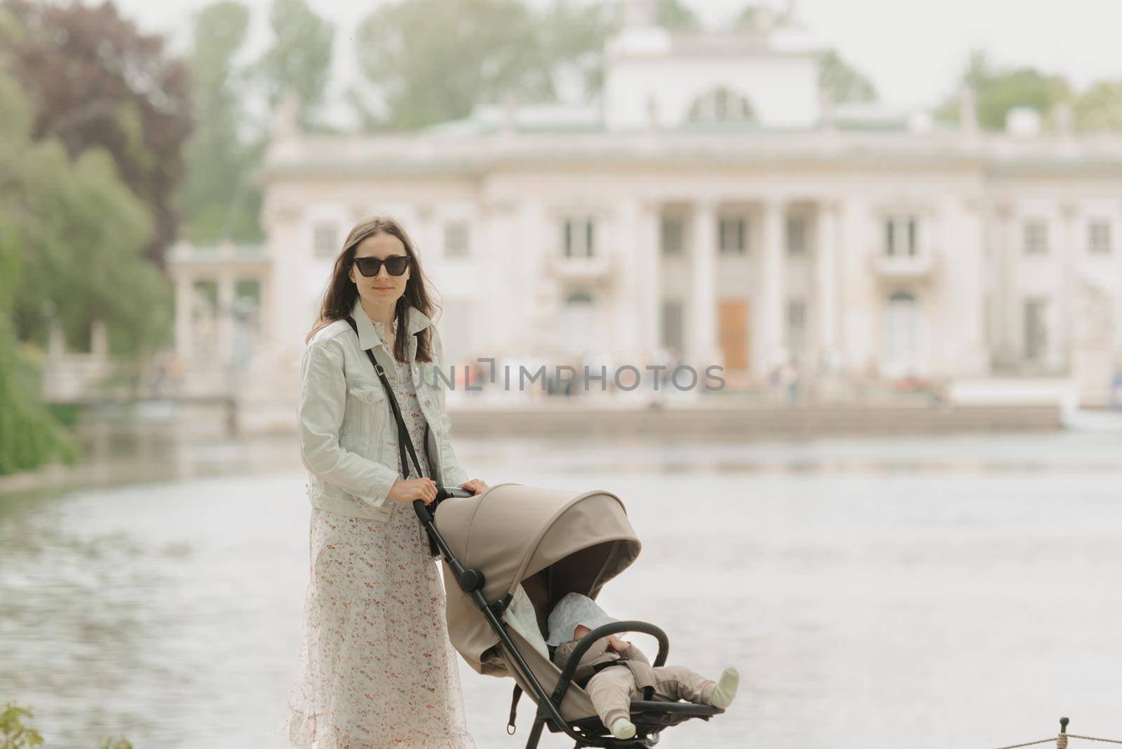 A mother is posing with her baby in the stroller on the territory of the palace. A mom in sunglasses with a baby in the carriage with classical architecture in the background in the park.