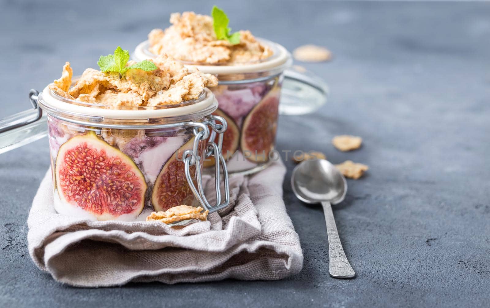 Homemade healthy yoghurt in glass pot with cereals, figs, mint on rustic concrete background. Healthy morning breakfast. Freshly made yoghurt with oat granola and fruit. Yoghurt in jar with muesli