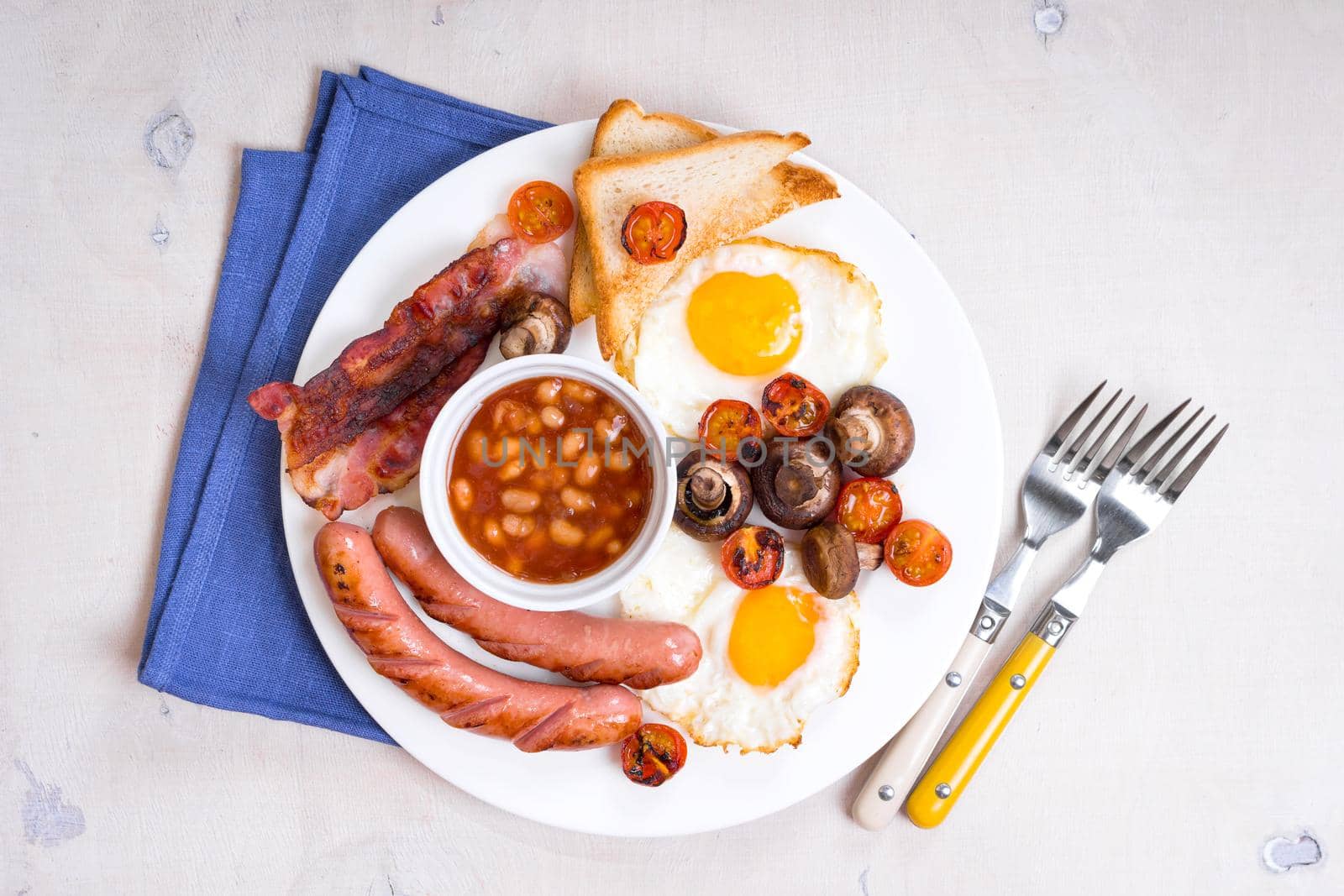 Full english breakfast with fried eggs, tomatoes, sausages, bacon, mushrooms, toasts and beans. Breakfast on a white plate with forks and blue napkin on a white wooden table. Top view