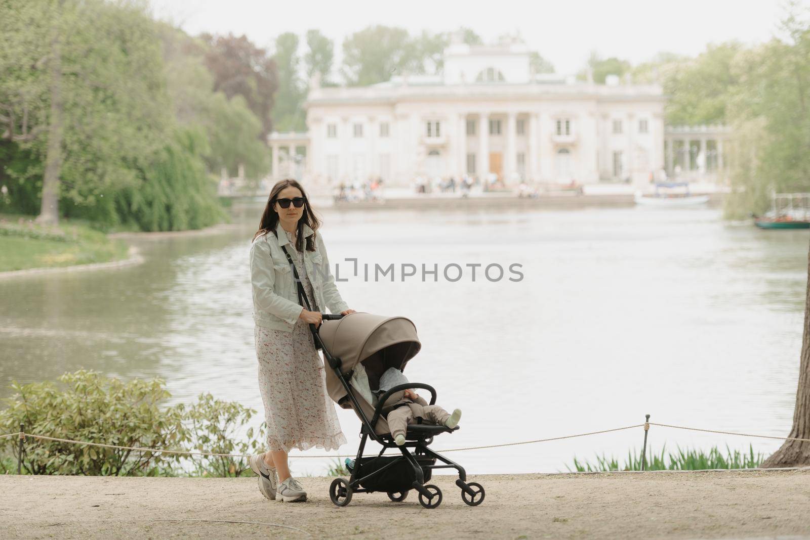 A mother in full growth is posing with her baby in the stroller on the territory of the palace. A mom with a baby in the carriage with classical architecture in the background in the park.