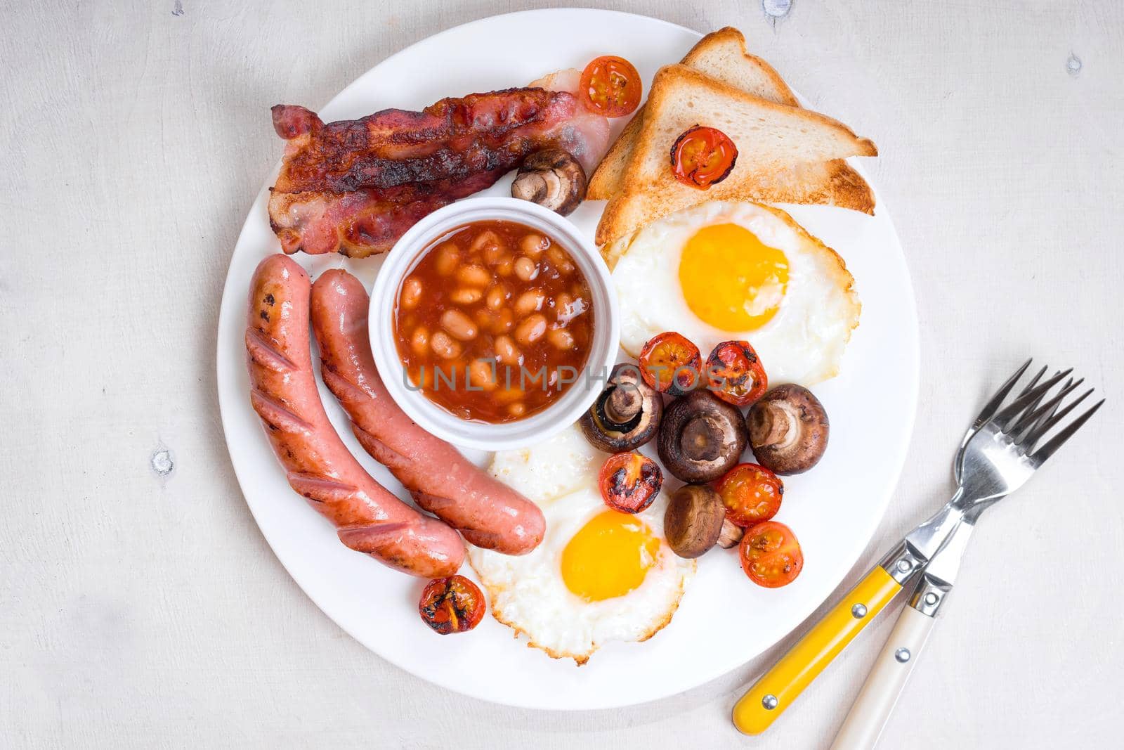 Full english breakfast with fried eggs, tomatoes, sausages, bacon, mushrooms, toasts and beans. Breakfast on a white plate with forks on the white wooden table. Top view