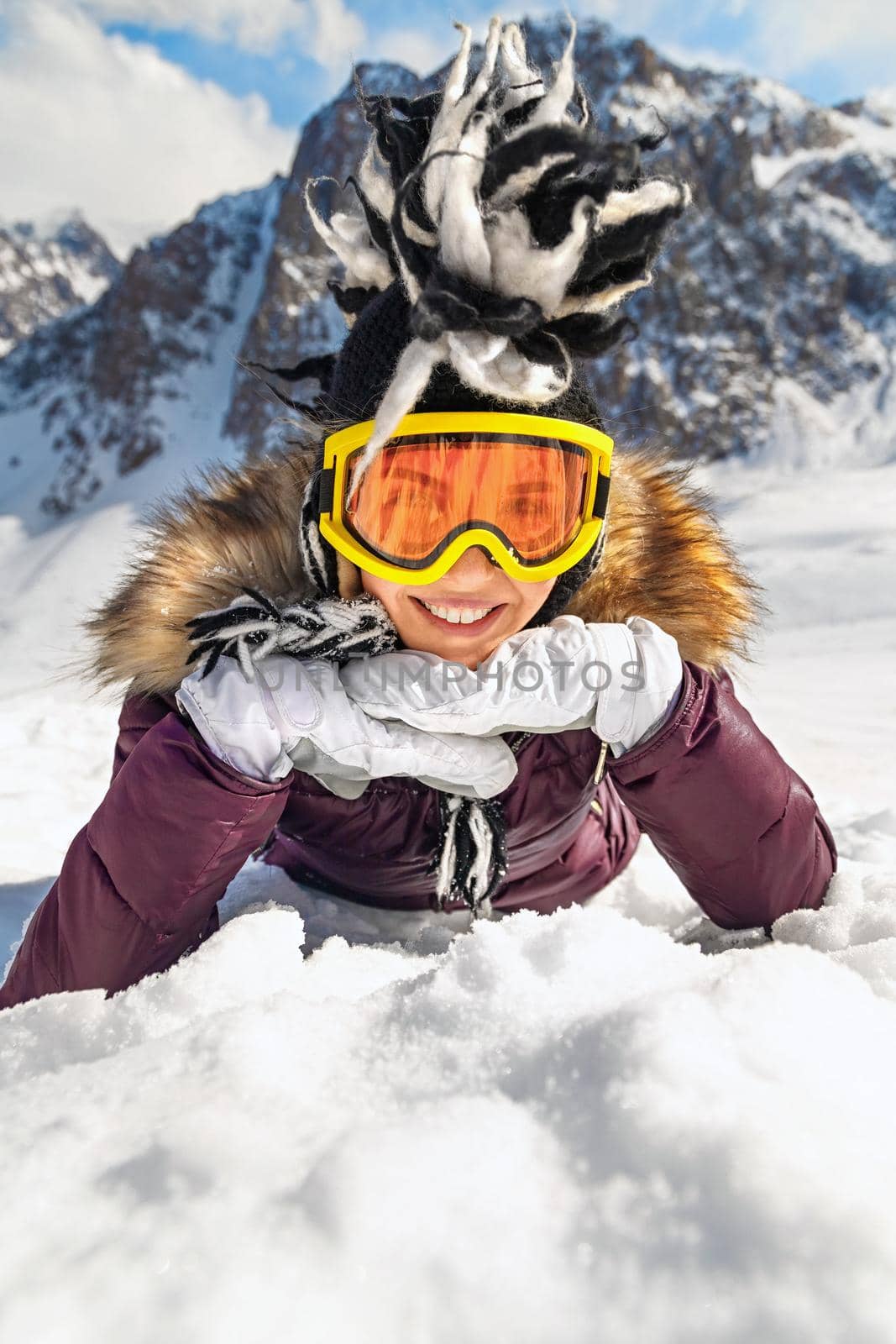 Portrait of playful hipster woman lying on snow at mountain in sunny day with blue sky and big rock in background. by Rom4ek