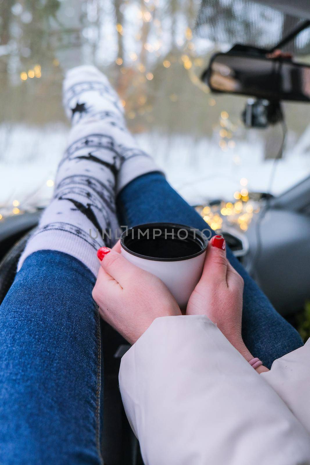 Woman legs with socks hand holding thermos cup of coffee or tea in winter car. Christmas socks. Garland defocused lights by anna_stasiia