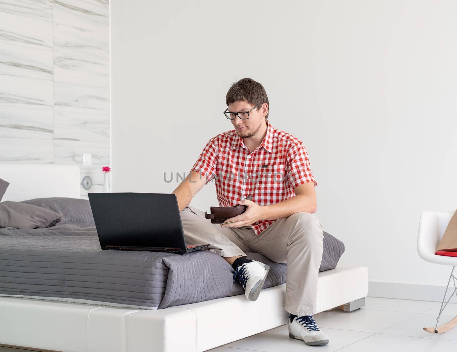 Online shopping concept. Young man sitting on the bed and shopping online using tablet holding a wallet, copy space
