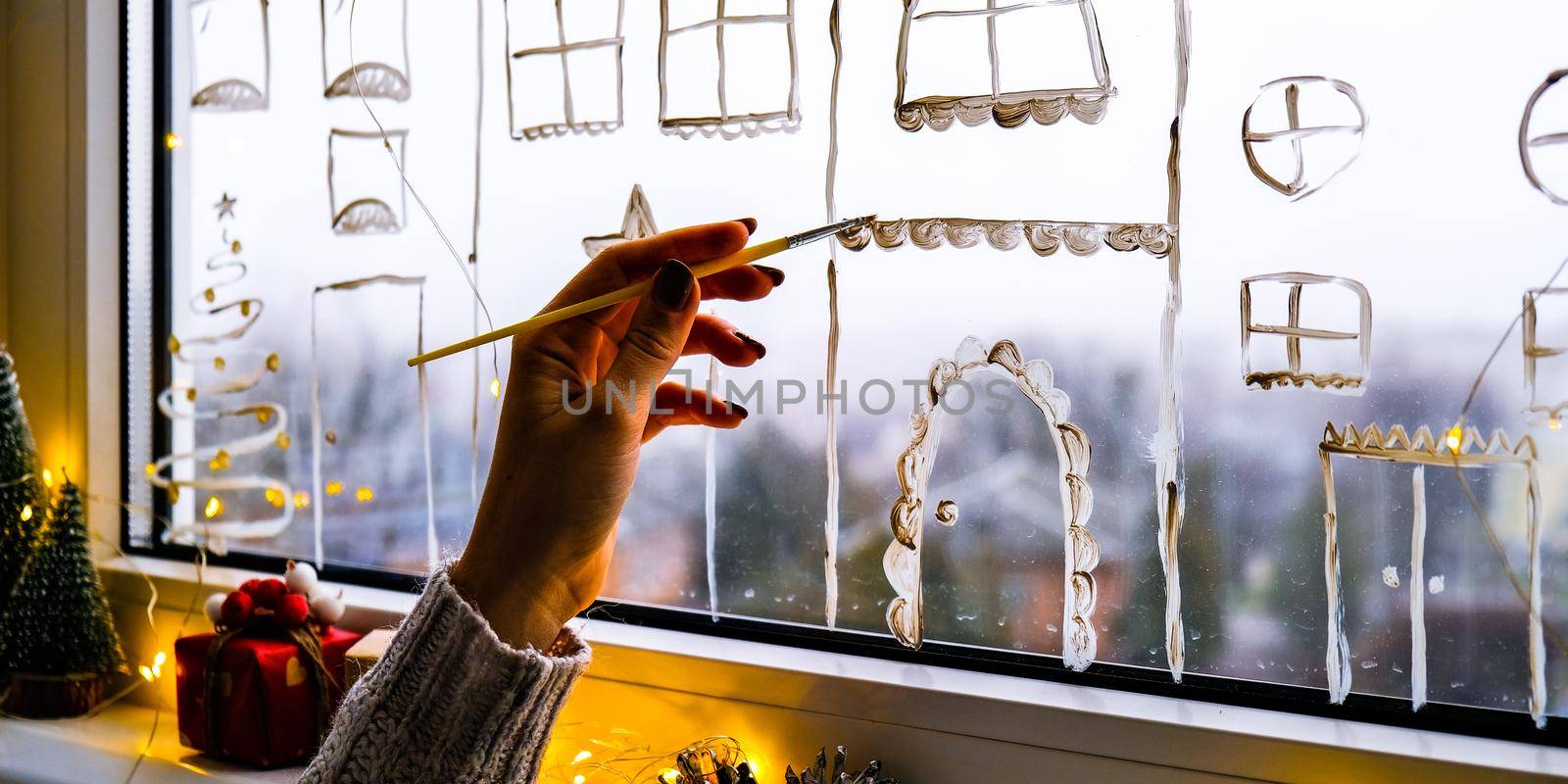 Female hand draws christmas holidays decoration painted on window glass. New Years scenery. city buildings garlands lights. hygge, decoration and christmas concept. festive garland string at home