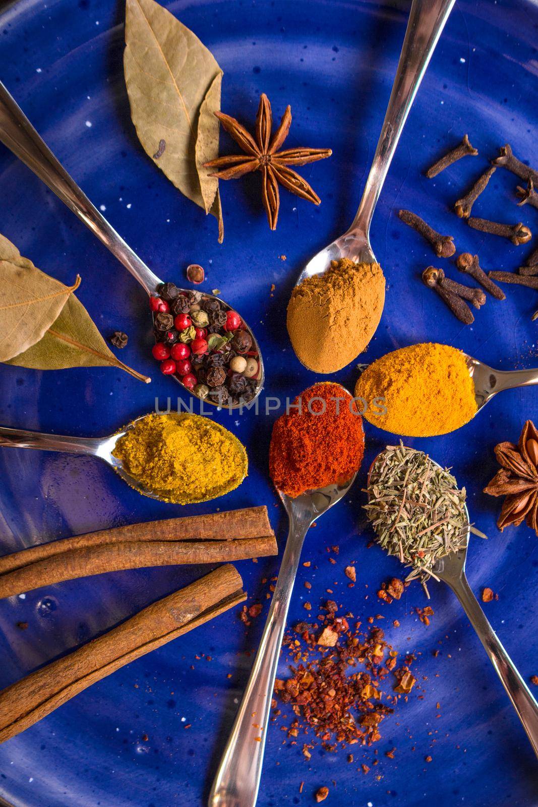 Different colorful spices in little spoons on dark blue background. Curry, turmeric, cinnamon sticks, bay leaves, sumac, cloves, peppercorn, paprika, rosemary, star anise. Top view. Closeup