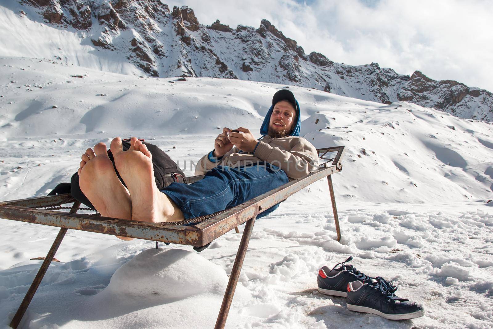 A man in winter with barefoot feet is resting on an iron bed in the mountains. Winter vacation fun concept