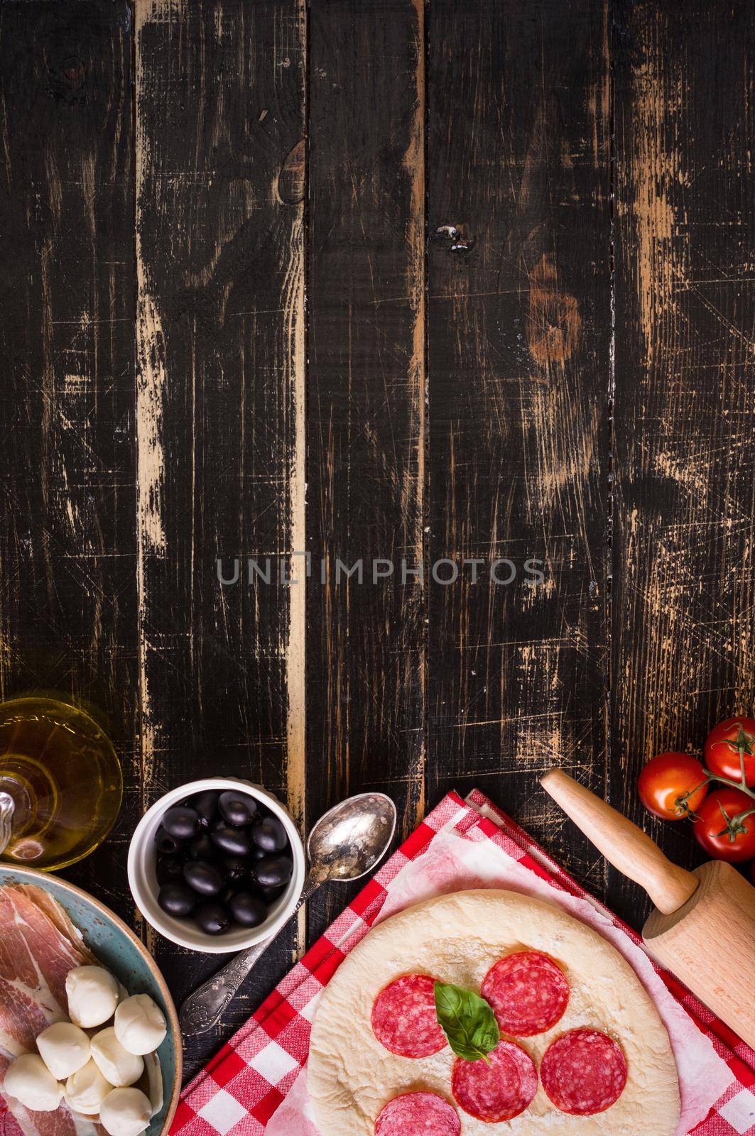 Pizza making background. Ingredients for making pizza. Space for text. Pizza dough, flour, cheese, mozzarella, tomatoes, basil, pepperoni, olives and rolling pin over black wooden background. Top view