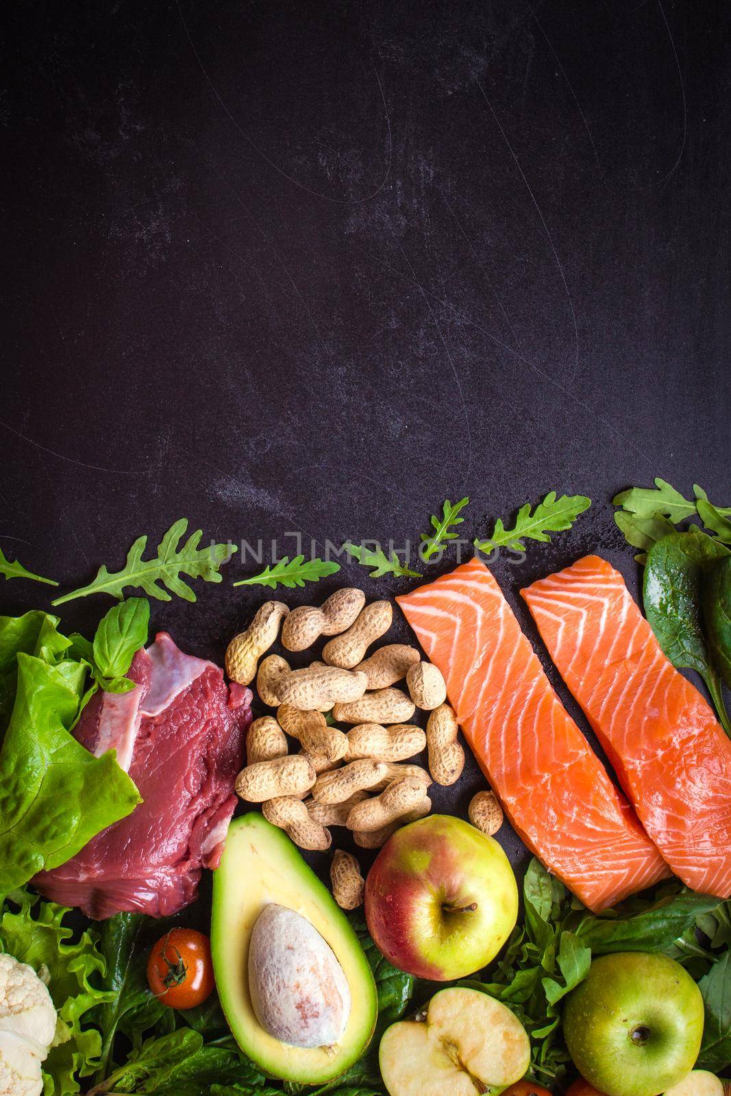 Fresh vegetables, fruits, fish, meat, nuts on black chalk board background. Сauliflower, avocado, apples, tomatoes, salmon, beef, spinach, herbs. Diet/healthy/paleo food. Ingredients. Space for text