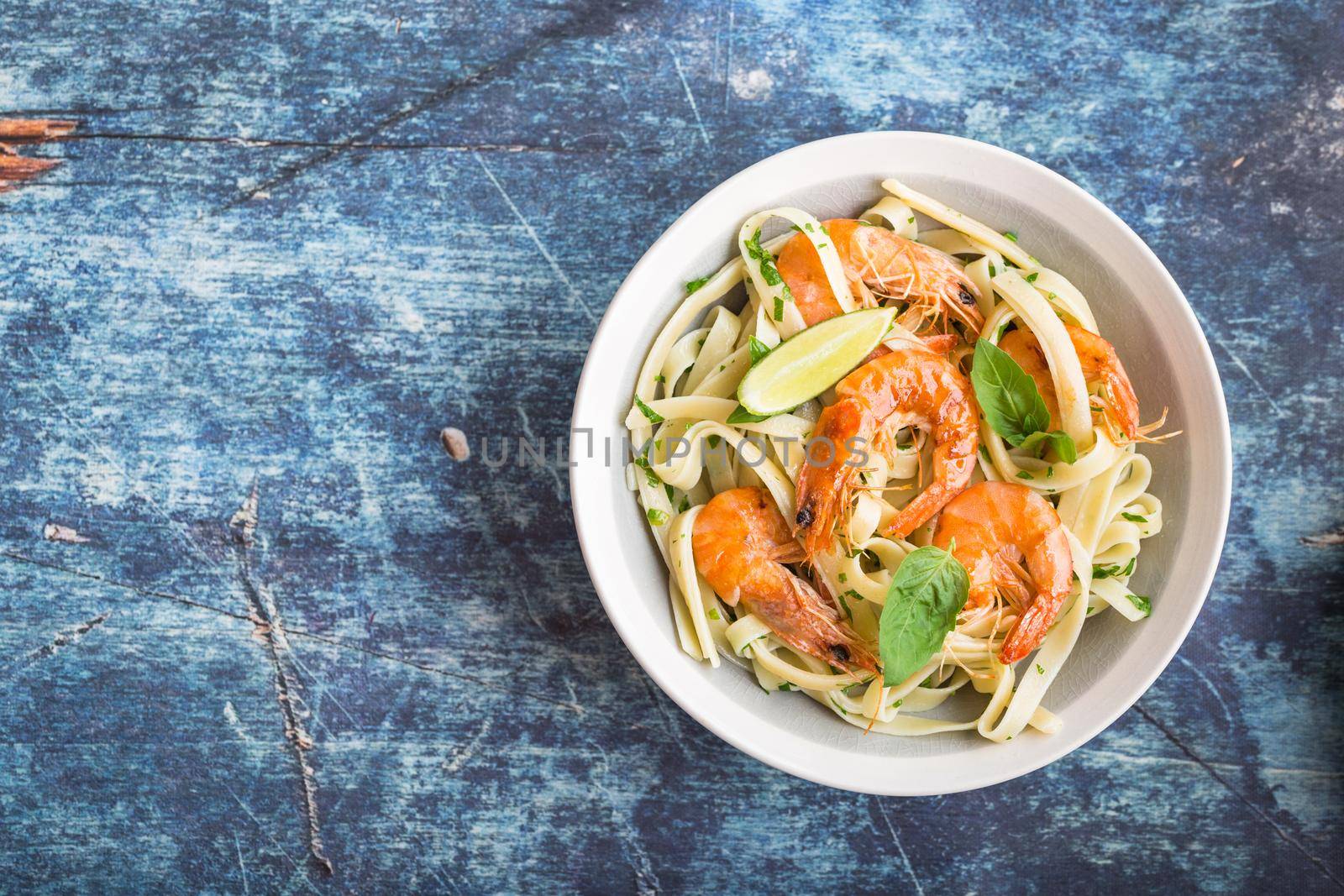 Homemade pasta with fried shrimps and basil on rustic wooden blue background. Space for text. Pasta with seafood in plate. Top view. Closeup. Mediterranean cuisine. Pasta for dinner or lunch. Overhead