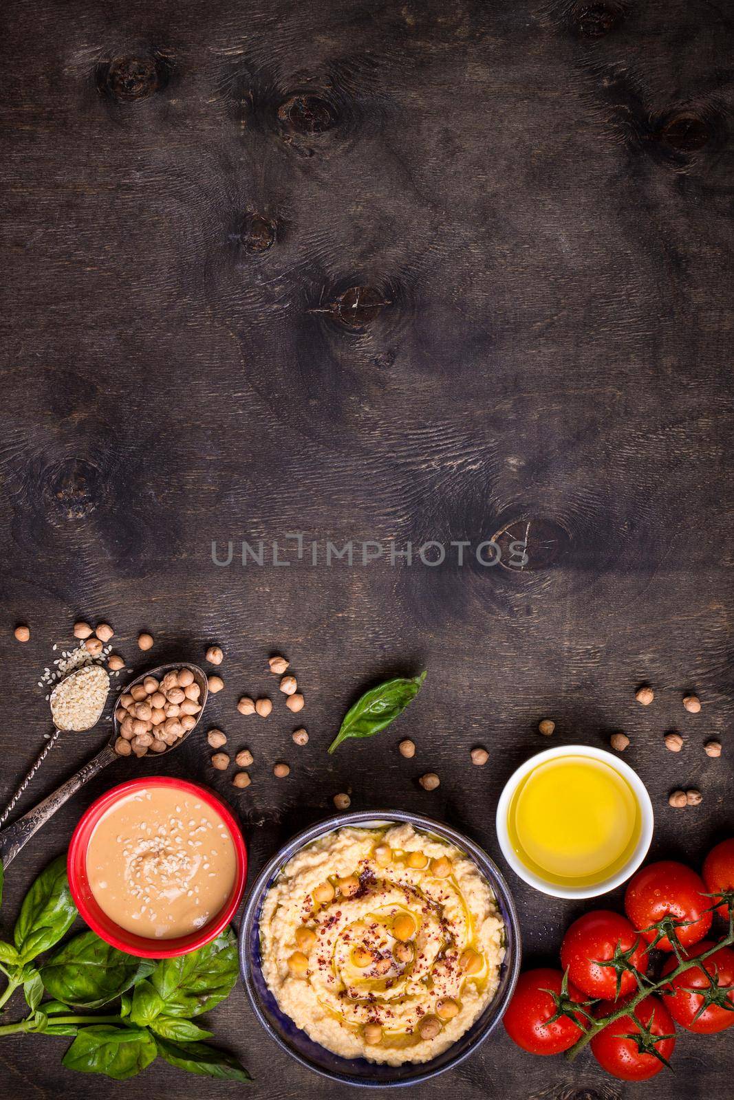 Bowl with hummus, chickpea, tahini, olive oil, sesame seeds, cherry tomatoes and herbs on dark rustic wooden background. Space for text. Food frame. Middle eastern cuisine. Top view. Hummus background