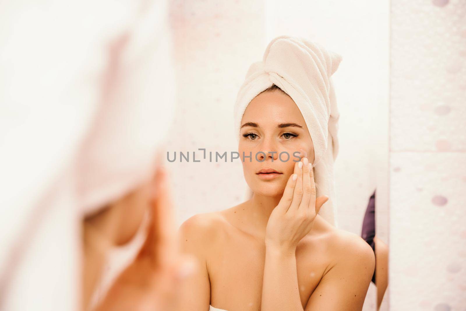 Young beautiful woman using face cream moisturizing lotion after bath. Pretty attractive girl in a towel on her head stands in front of a mirror in a home bathroom. Daily hygiene and skin care by Matiunina