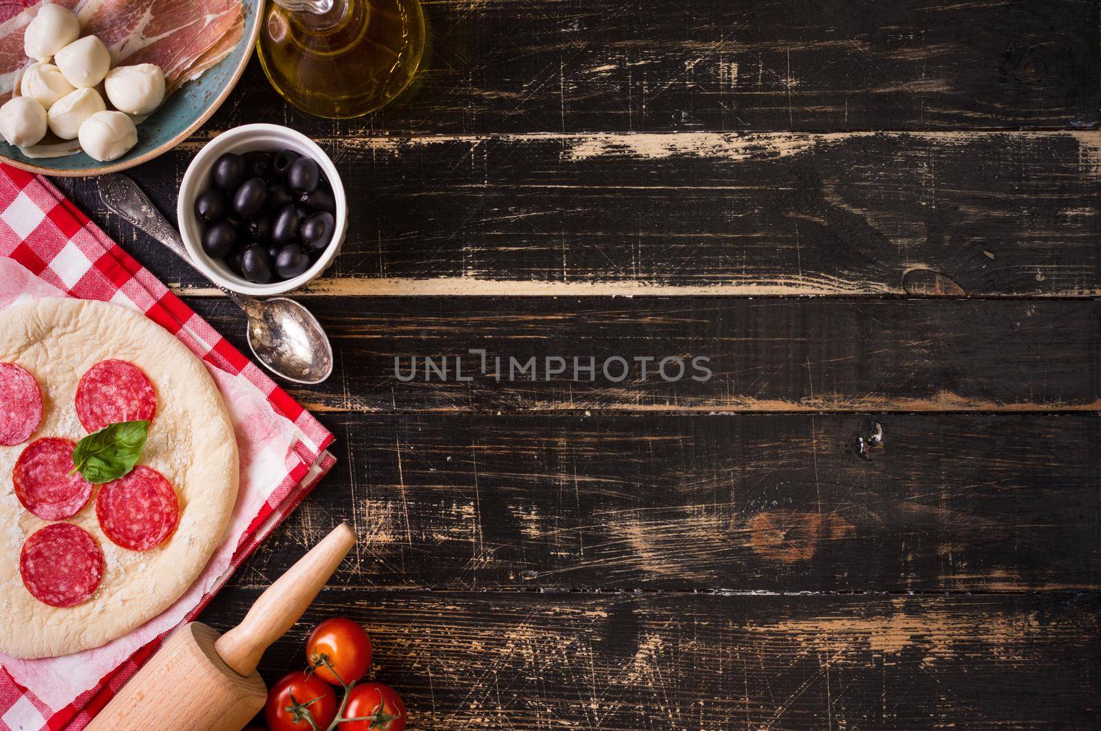 Pizza making background. Ingredients for making pizza. Space for text. Pizza dough, flour, cheese, mozzarella, tomatoes, basil, pepperoni, olives and rolling pin over black wooden background. Top view