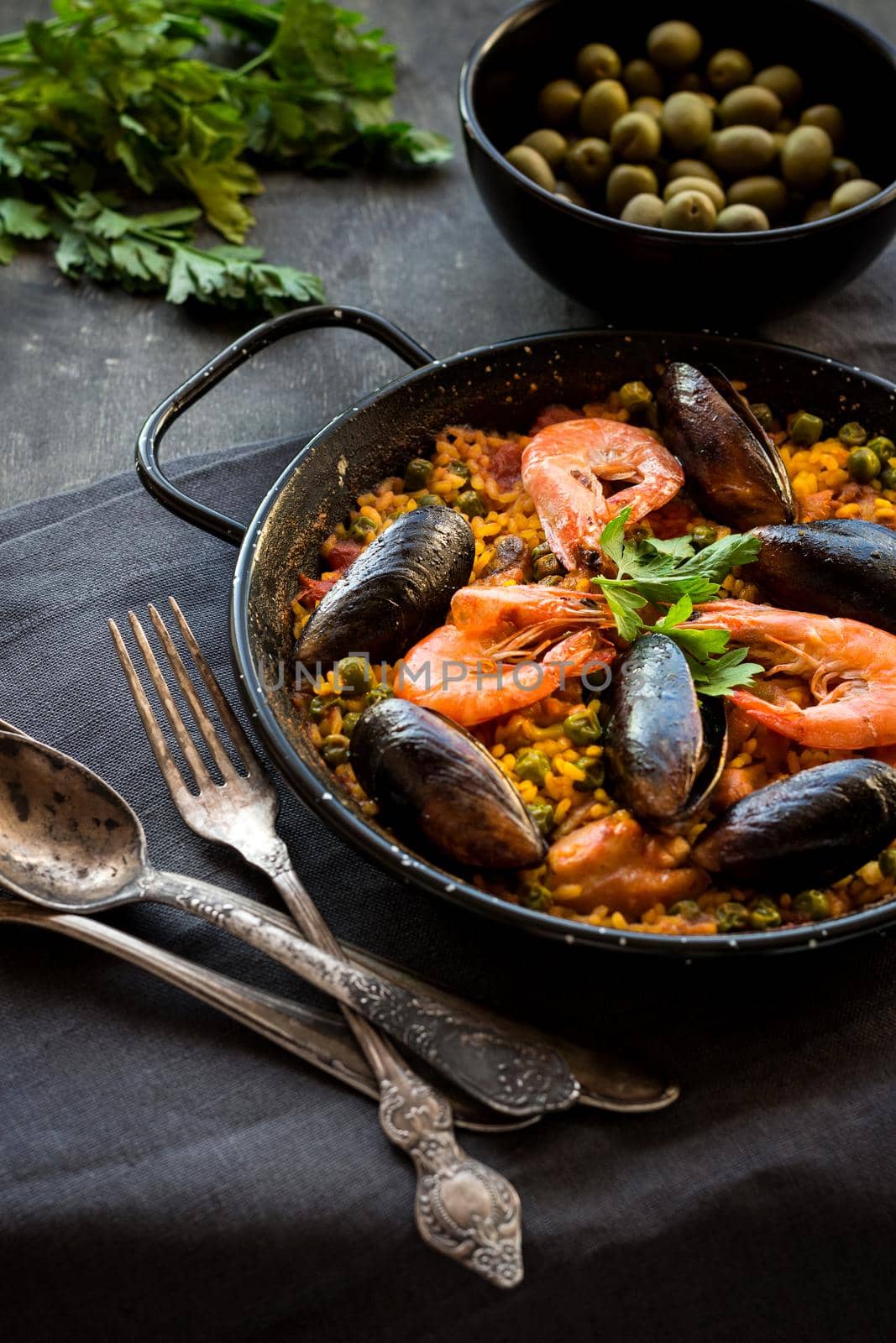 Paella in black pan with rice, shrimps, mussels, squid and meat, bowl with olives and vintage cutlery. Seafood paella, traditional spanish dish. Paella on rustic black wooden table. Selective focus