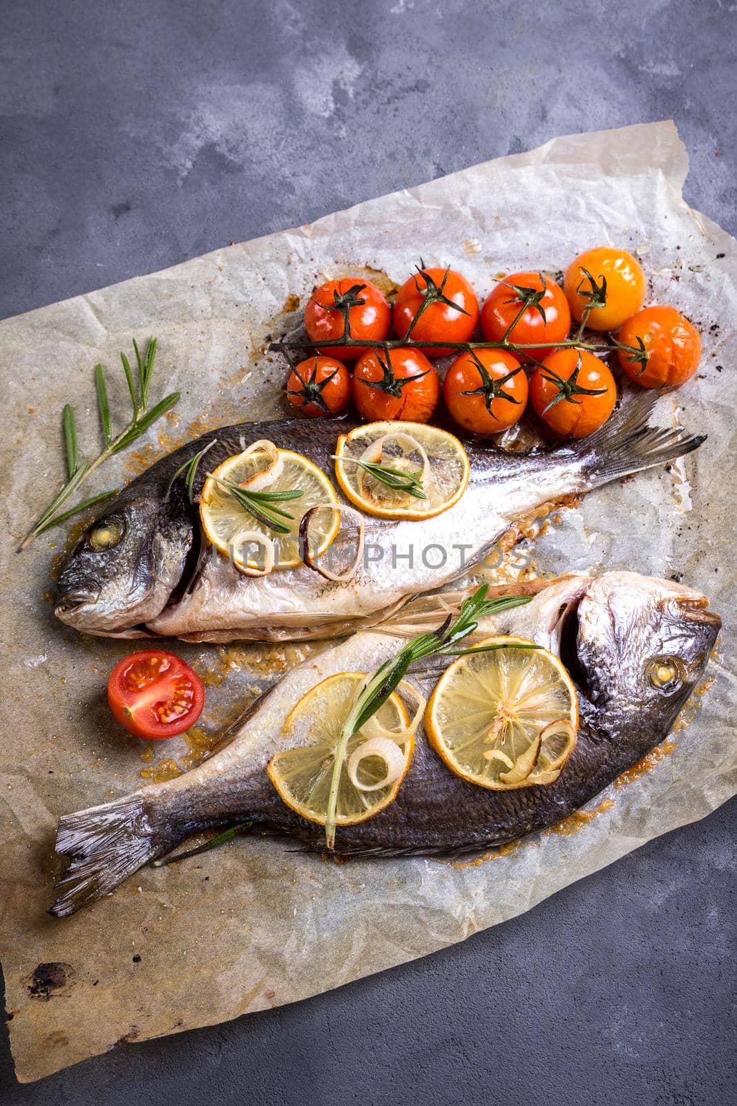 Tasty baked whole fish on baking paper. Baked sea bream with lemon, onion, herbs, cherry tomatoes, spices on dark rustic background. Grilled delicious fish. Diet and healthy food. Top view