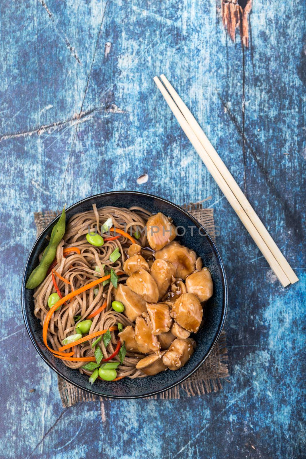 Asian noodles with chicken, vegetables, bowl, rustic wooden blue background. Closeup. Top view. Soba noodles, teriyaki chicken, edamame, chopsticks. Asian style dinner/lunch. Chinese/Japanese noodles