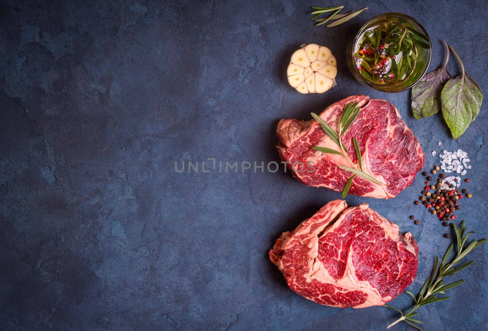 Raw juicy ribeye steaks with seasonings ready for roasting on rustic concrete background. Fresh marbled meat steaks with herbs, garlic, olive oil, whole pepper and rock salt. Space for text. Top view