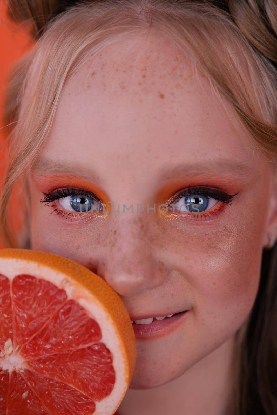 young girl with grapefruit slice on orange background. curly hair and two ponytails.