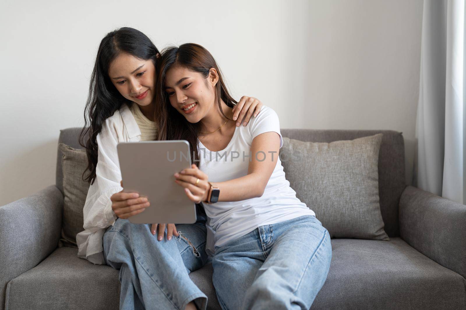 Happy young lesbian couple hugging having fun using digital tablet relaxing on couch at home. Two smiling asian women friends holding computer looking at screen enjoying surfing online watching videos.