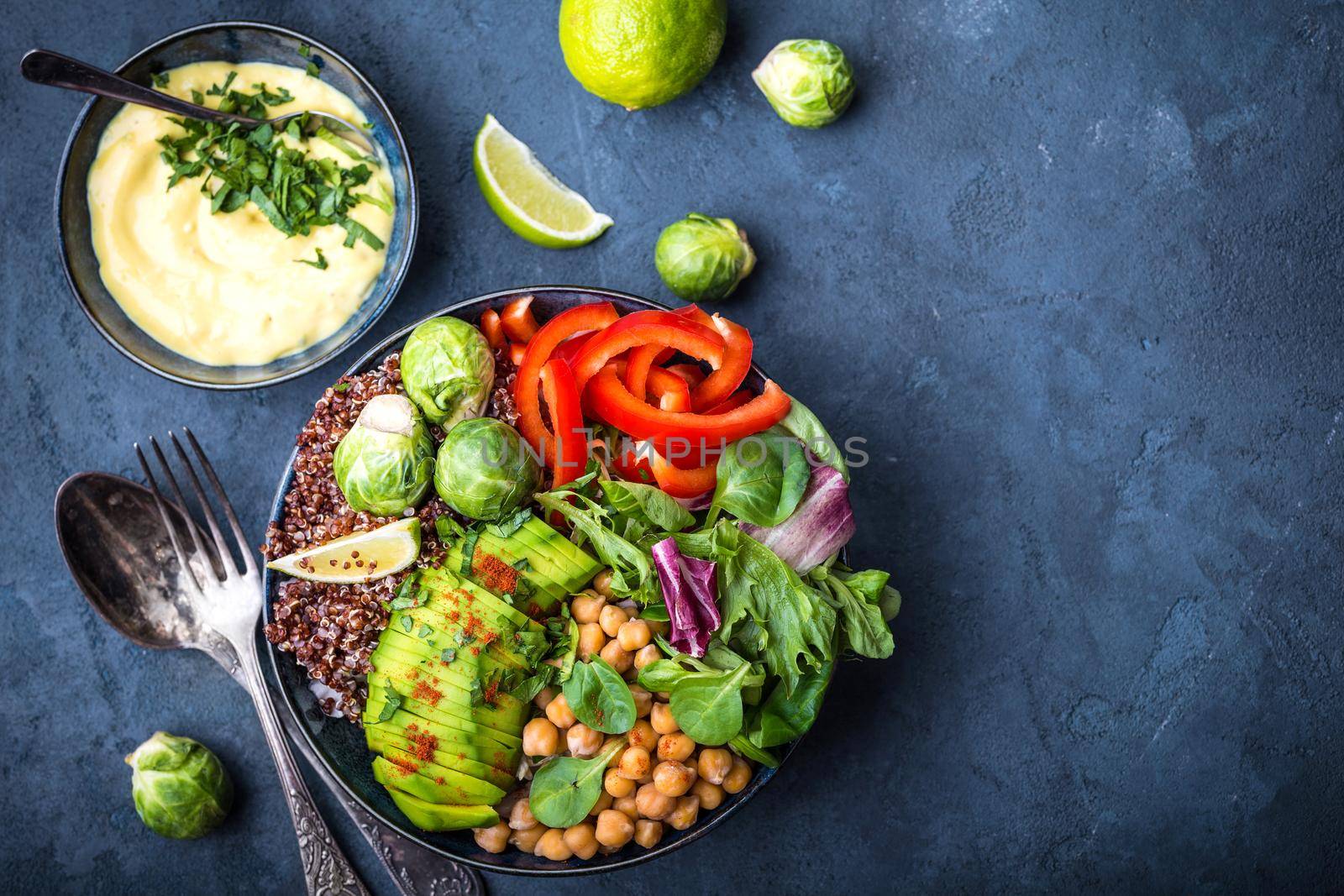 Bowl with healthy salad, dip, blue stone background. Top view. Space for text. Buddha bowl with chickpea, avocado, quinoa seeds, red bell pepper, fresh spinach, brussels sprout, lime. Vegetarian salad