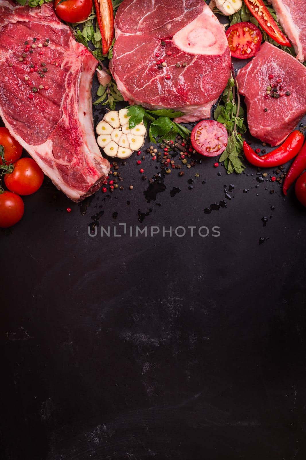 Raw juicy meat steaks ready for roasting on a black chalk board background. Rib eye steak on the bone, veal shank (ossobuco), fillet with cherry tomatoes, hot pepper and herbs. Space for text