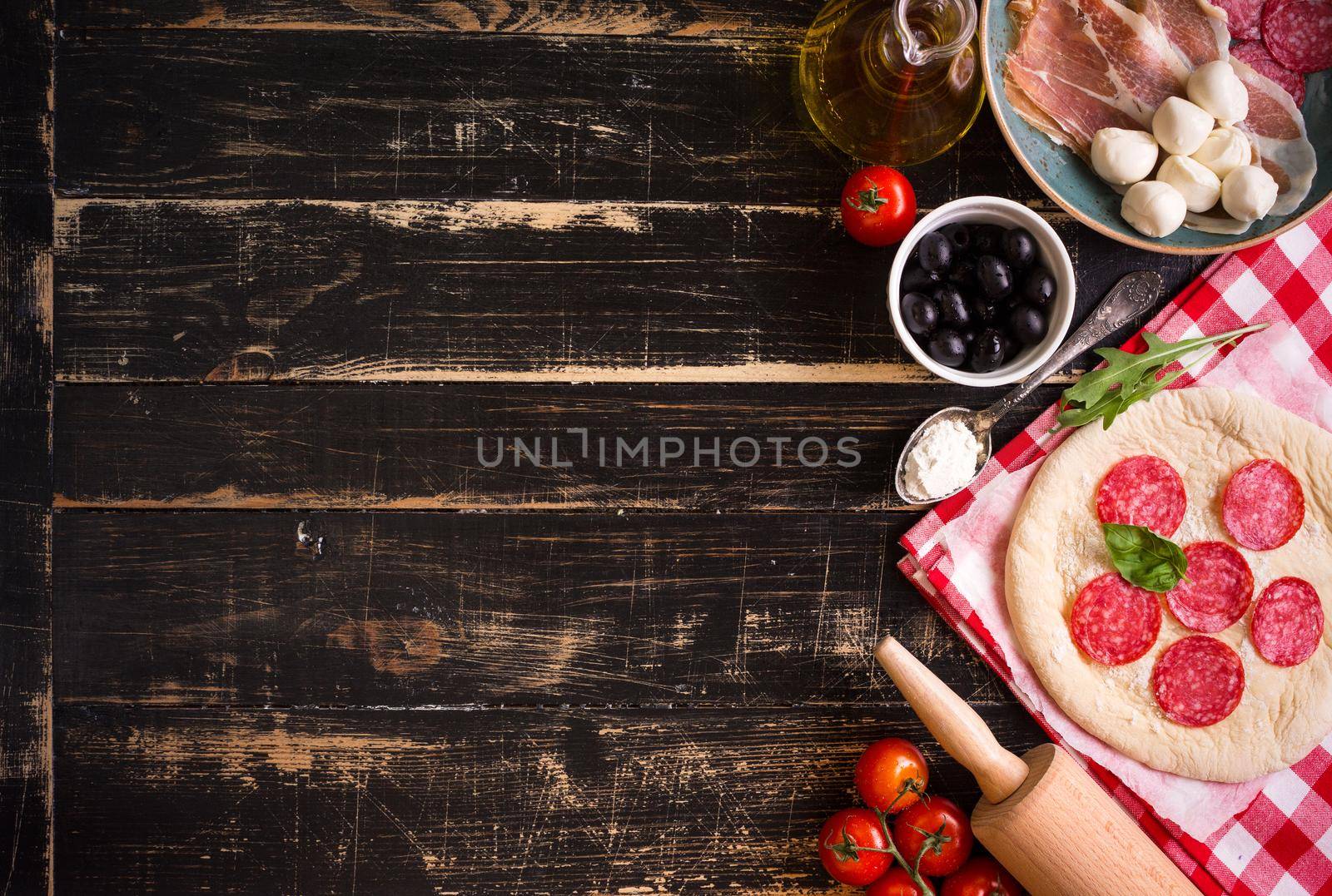 Pizza making background. Ingredients for making pizza. Space for text. Pizza dough, flour, cheese, mozzarella, tomatoes, basil, pepperoni, olives and rolling pin over black wooden background. Top view