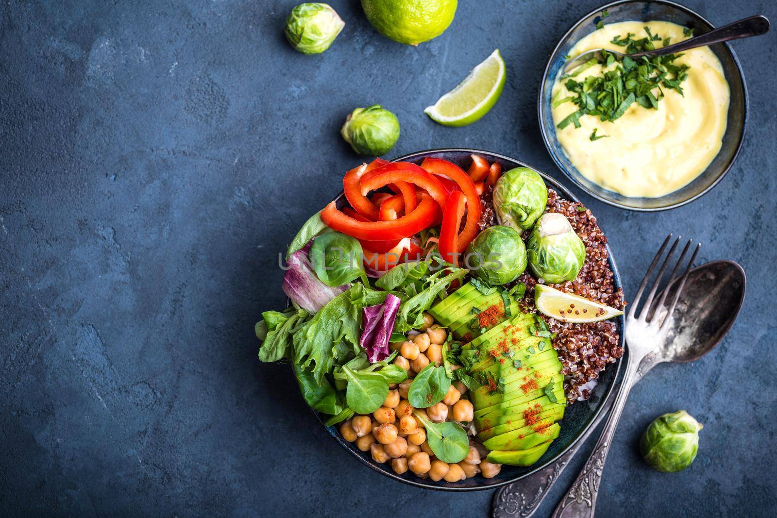 Bowl with healthy salad, dip, blue stone background. Top view. Space for text. Buddha bowl with chickpea, avocado, quinoa seeds, red bell pepper, fresh spinach, brussels sprout, lime. Vegetarian salad