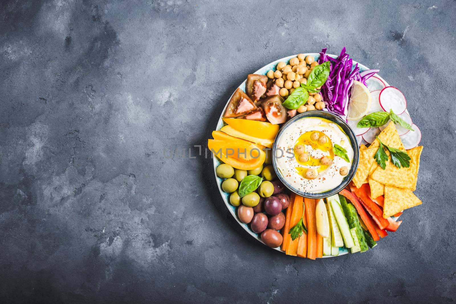 Hummus platter with assorted snacks, stone background. Space for text. Hummus, vegetables sticks, chickpeas, olives, pita chips. Plate, Middle Eastern/Mediterranean meze. Party/finger food. Top view