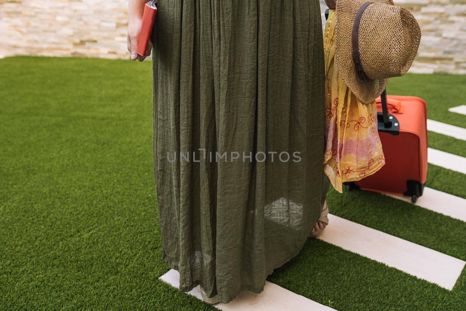 cropped shot of a young woman, entering the hotel reception with her suitcase on wheels. woman on a pleasure trip dressed in summer clothes. Outside, dim sunlight, grass floor with white tiles.