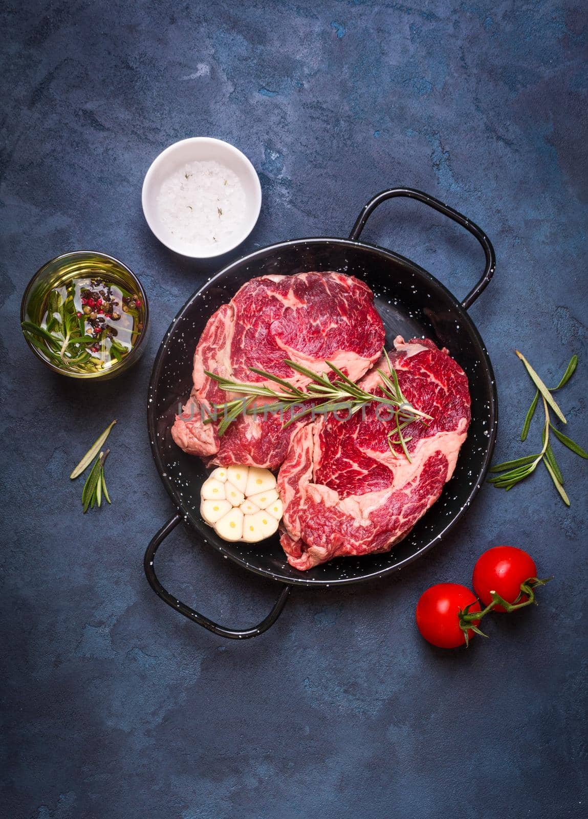 Raw juicy steaks with seasonings in a black pan ready for roasting on rustic concrete background. Fresh marbled meat steaks with herbs, garlic, olive oil, pepper, salt and tomatoes. Top view