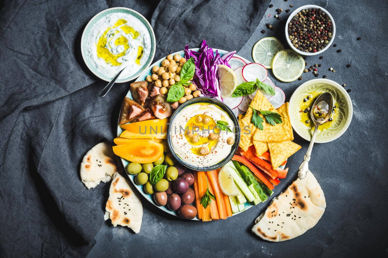 Meze platter with hummus, yoghurt dip, assorted snacks. Hummus in bowl, vegetables sticks, chickpeas, olives, pita, chips. Plate with Middle Eastern/Mediterranean meze. Party/finger food. Top view