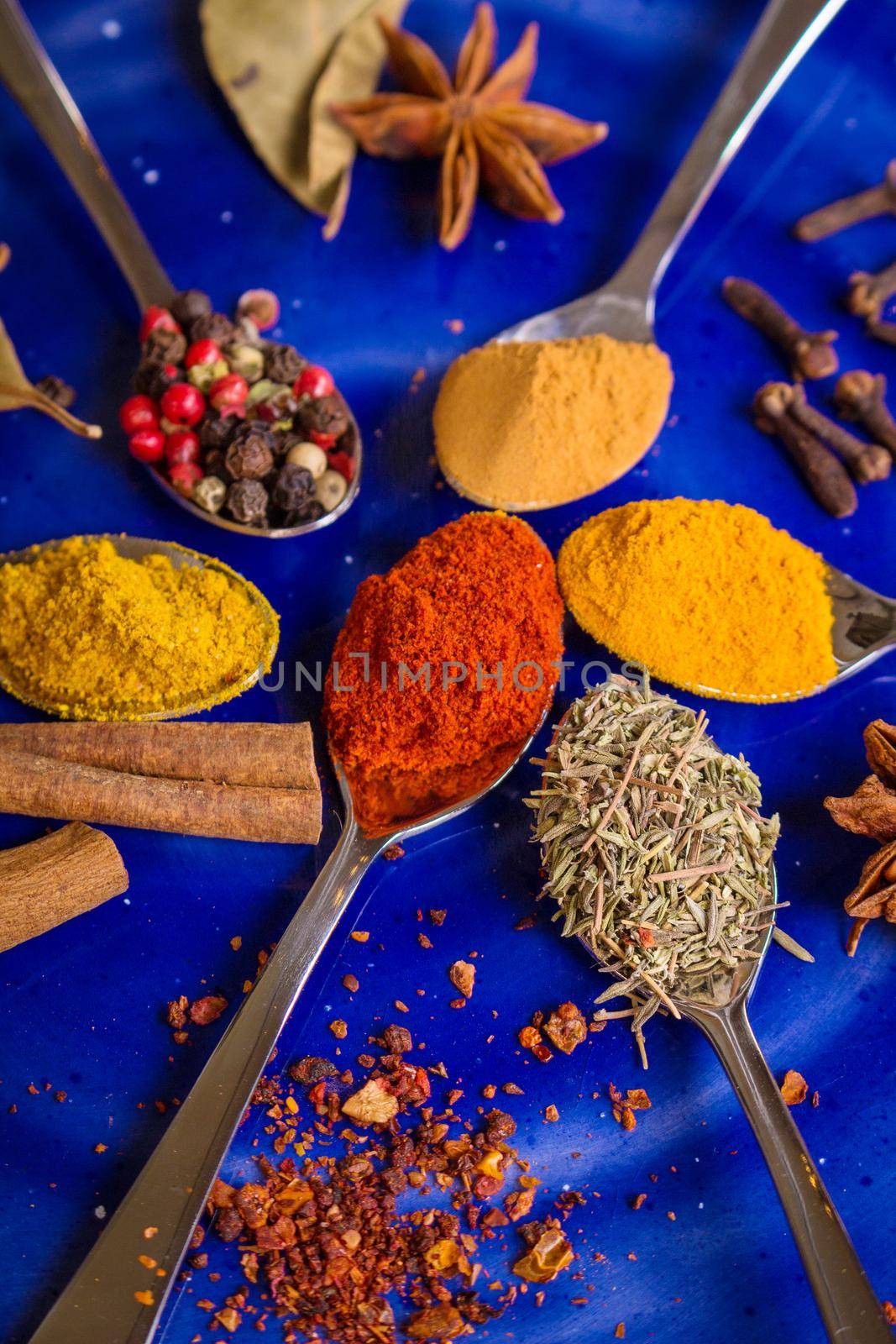 Different colorful spices in little spoons on dark blue background. Curry, turmeric, cinnamon sticks, bay leaves, sumac, cloves, peppercorn, paprika, rosemary, star anise. Top view. Closeup