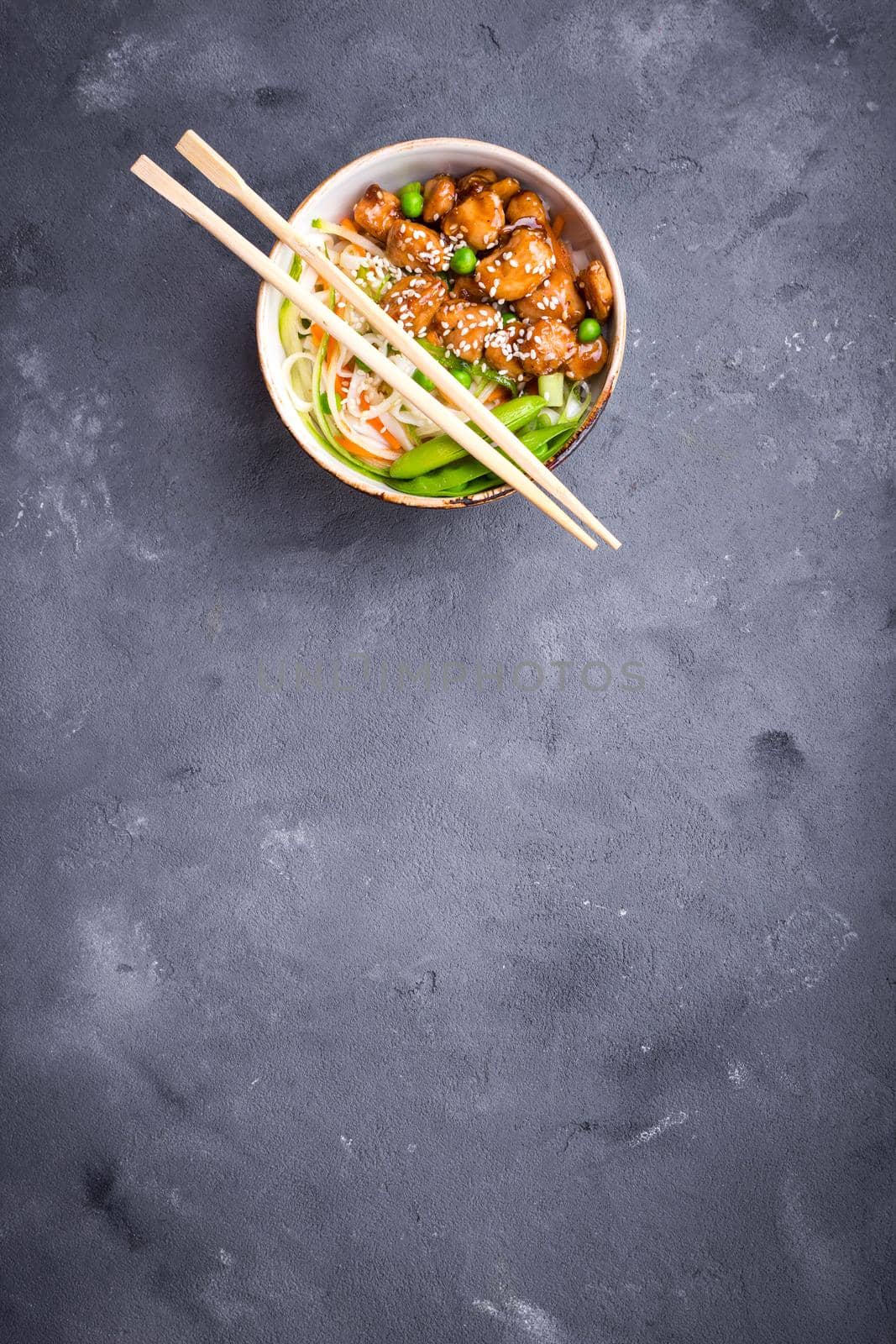 Asian style noodles with teriyaki chicken, vegetables and green peas pods. Noodles in bowl on rustic concrete background. Chinese/thai/japanese style stir fry noodles. Space for text. Top view