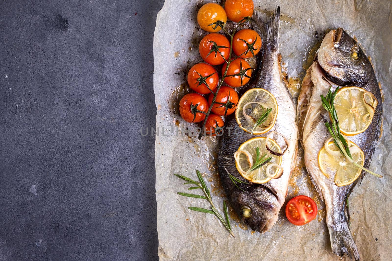 Tasty baked whole fish on baking paper. Baked sea bream with lemon, onion, herbs, cherry tomatoes, spices on dark rustic background. Grilled delicious fish. Diet and healthy food. Top view