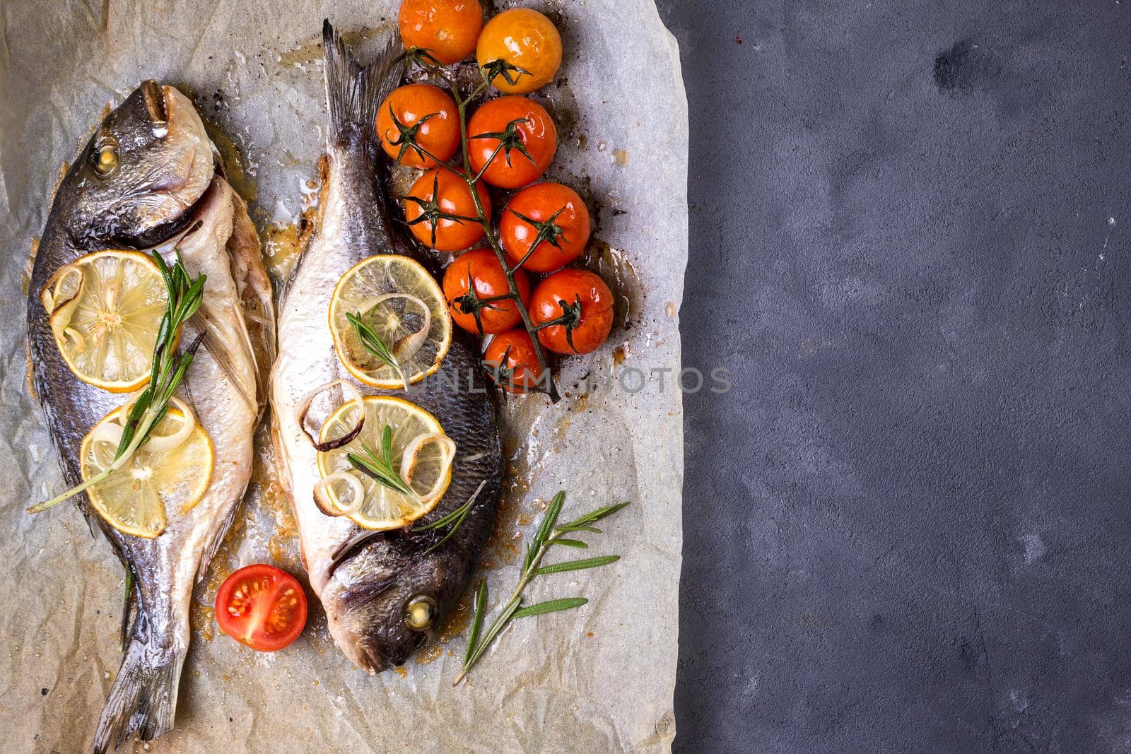 Tasty baked whole fish on baking paper. Baked sea bream with lemon, onion, herbs, cherry tomatoes, spices on dark rustic background. Grilled delicious fish. Diet and healthy food. Top view